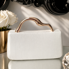 A white textured leather clutch bag with a wavy rose gold metal handle, displayed on a reflective surface with a white rose in a gold vase and a black decorative mirror in the background.
