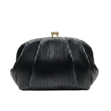 A hand with long manicured nails holds a black pleated velvet clutch bag with a gold-tone metal clasp against a warm beige background with vertical light strips.