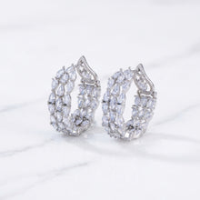 A pair of large silver teardrop-shaped hoop earrings paved with clear marquise and round stones, displayed on a beige jewelry box with a blurred floral background.