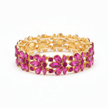 A gold-plated stretch bracelet featuring a row of red faceted gemstones in a floral pattern, resting on a light-colored surface.