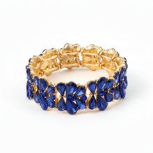 A gold-plated stretch bracelet featuring a row of dark blue faceted rhinestones in a floral pattern, resting on a light-colored surface.