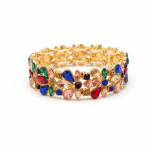 A gold-plated stretch bracelet adorned with colorful rhinestones in red, green, blue, and black, displayed on a light-colored surface.