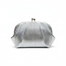 A hand holding a silver metallic pleated clutch bag with a double-bell clasp, set against a dark brown background with vertical gold light strips.