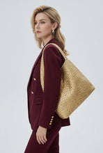 A woman in a black blazer carries a large woven straw tote bag over her shoulder, photographed against a clean light gray studio background.