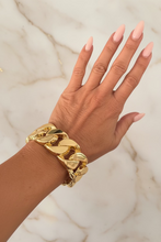 A close-up of a person's hand and wrist wearing a thick, chunky gold Cuban link chain bracelet, resting against a dark marble surface.