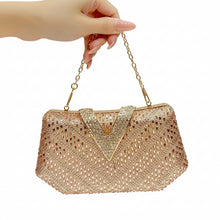 A hand with long red manicured nails holds a sparkling rhinestone evening handbag with a gold chain strap. The bag features a textured, rhinestone-covered body with a prominent V-shaped crystal detail on the front flap, set against a warm, dark brown background with vertical gold light bars.