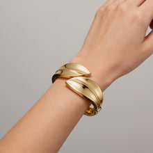 A close-up of a person's wrist wearing a polished gold leaf-shaped cuff bracelet, set against a dark textured background with vintage pocket watch props.