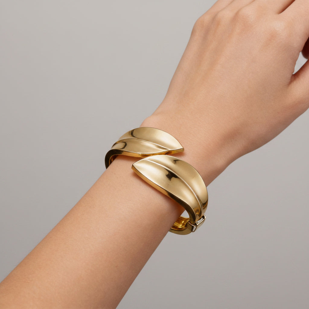 A close-up of a person's wrist wearing a polished gold leaf-shaped cuff bracelet, set against a dark textured background with vintage pocket watch props.