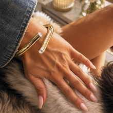 A close-up lifestyle shot of a person's hand resting on a fluffy faux fur surface, wearing a chunky gold cuff bracelet with a clear rhinestone. The person is also wearing a blue denim sleeve, and a lit candle and flowers are visible in the softly blurred background.