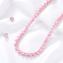 A heart-shaped crystal necklace in a rose gold setting, displayed on a black fabric background alongside matching heart stud earrings on a dark blue cloth.