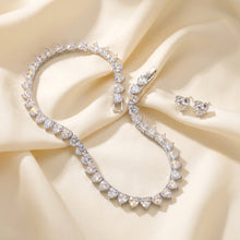 A silver-toned jewelry set featuring a heart-link bracelet and matching stud earrings, both adorned with clear heart-shaped gemstones, displayed on a dark navy fabric background.
