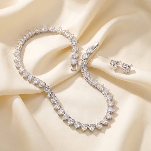 A silver-toned jewelry set featuring a heart-link bracelet and matching stud earrings, both adorned with clear heart-shaped gemstones, displayed on a dark navy fabric background.